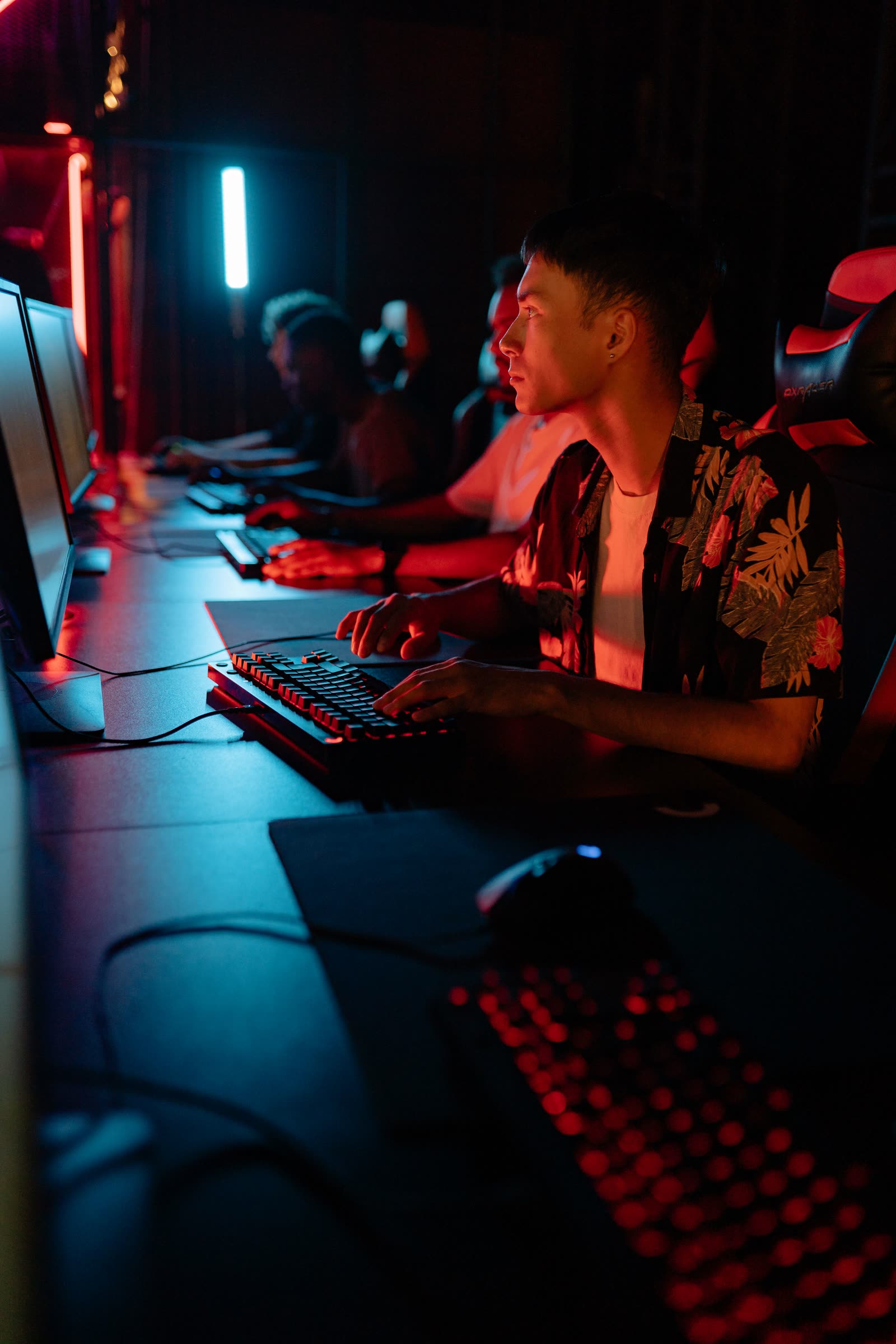 A focused player at a red-lit station in a row, hand on a mechanical keyboard, screen glowing in front